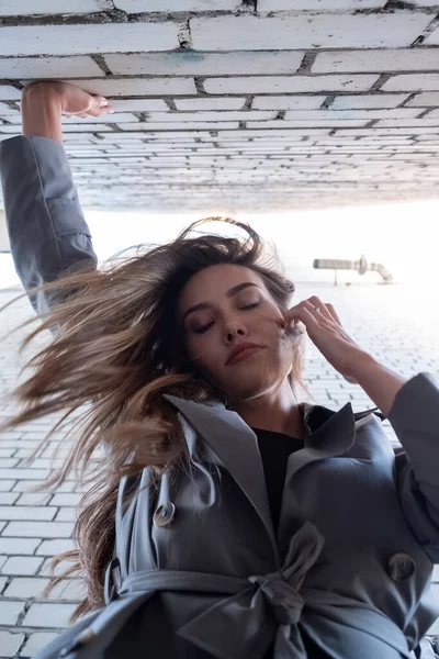 Trençkotlu Asyalı güzel manken ellerini duvara dayayıp aşağıya bakıyor.