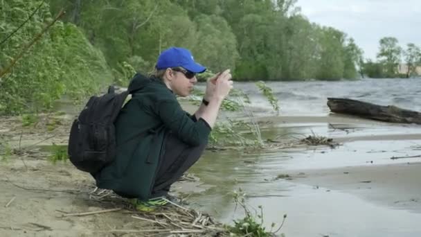 Jonge Kaukasische man zitten aan het strand van de rivier, met behulp van smartphone, het nemen van foto 's — Stockvideo