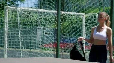 Spor kıyafetli sarışın genç bir kadın spor sahasındaki masaya sırt çantası bırakıyor.
