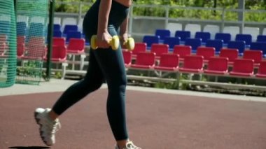 Genç formda sporcu kız spor sahasında dambıllarla hareket ediyor.