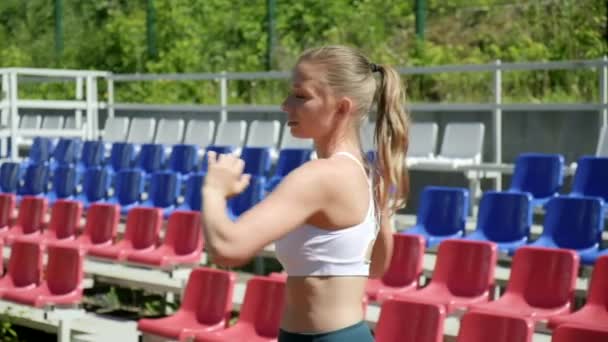 Young fit busty woman with ponytail in sportswear stretching at sports ground — Stockvideo