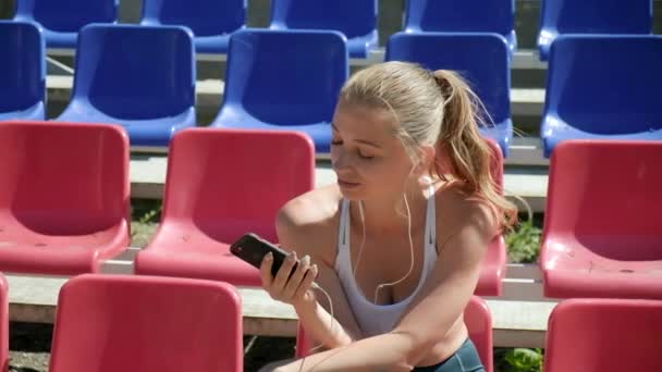 Ung kaukasisk kvinna med hörlurar lyssnar på musik på smartphone på stadion — Stockvideo