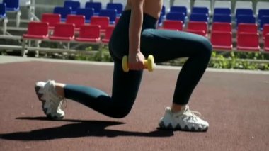 Genç formda sporcu kız spor sahasında dambıllarla hareket ediyor.