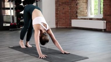 Genç, zayıf, beyaz bir kadın köpeğe doğru eğilip asana pozu veriyor.