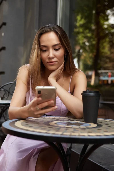 若いアジア系の女性がコーヒーを片手にテーブルに座りスマートフォンで — ストック写真