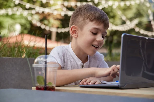 Kafenin verandasında oturmuş laptopla ders çalışan beyaz bir çocuk.