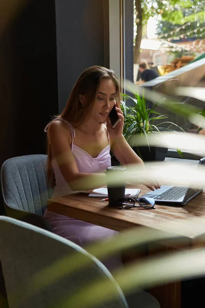 Νεαρή Ασιάτισσα που κάθεται σε καφετέρια με φορητό υπολογιστή, καλώντας από το smartphone — Φωτογραφία Αρχείου