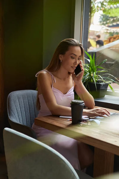 Νεαρή Ασιάτισσα που κάθεται σε καφετέρια με φορητό υπολογιστή, καλώντας από το smartphone — Φωτογραφία Αρχείου