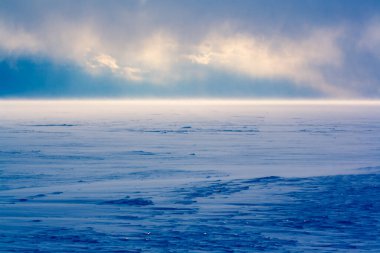 Baykal Gölü 'nde kar fırtınası. Güneş karların ve bulutların arasından parlıyor. Güçlü rüzgar karı süpürür. Maviyle boyanmış. Yatay.