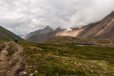 Turistler kasvetli havada dağlardaki taşlardan oluşan patika boyunca vadi boyunca yürürler. Küçük bir göl. Yüksek sıradağlar ve alçak bulutlar. Yatay.