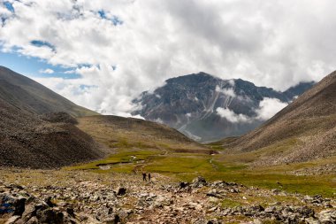 Dağlar arasında bir geçit olan dağ vadisi ve sonunda büyük bir dağ. Dağda alçak bulutlar. Birkaç turist patika boyunca gider. Dünyada bir sürü taş var. Yatay.