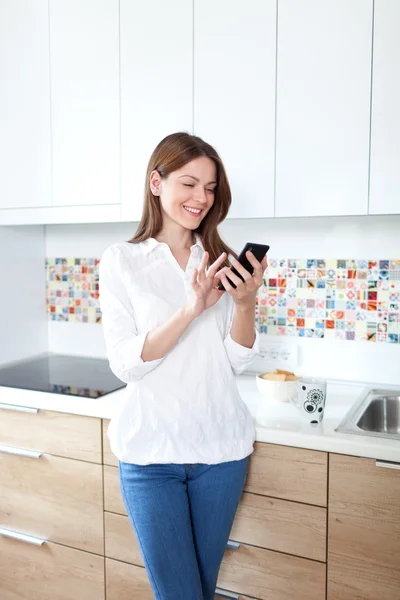 Ung kvinna med hjälp av mobiltelefon i köket — Stockfoto