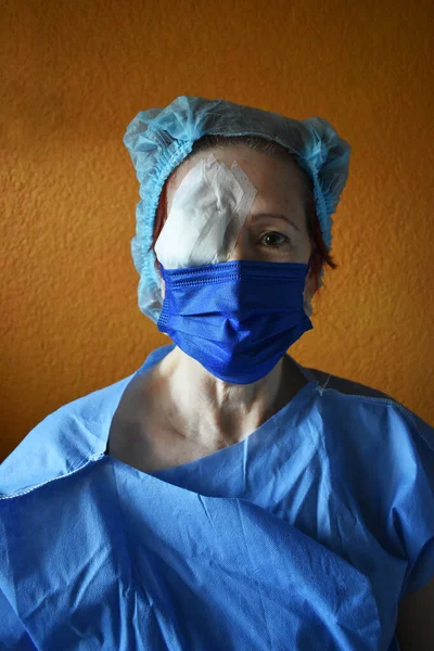 Woman operated on the right eye, wears a disposable gown and a disposable cap, brings the eye covered with a bandage, wears a blue mask