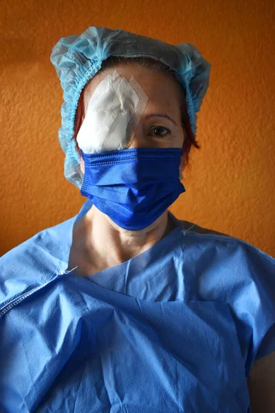 Woman operated on the right eye, wears a disposable gown and a disposable cap, brings the eye covered with a bandage, wears a blue mask