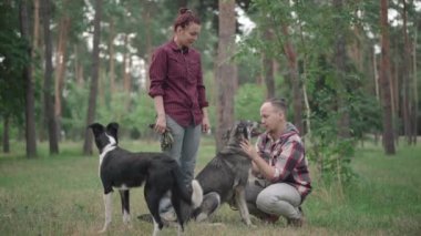 Ormanda duran kendine güvenen beyaz bir kadın ya da çöreklerin üzerine oturup köpekle konuşan pozitif bir adam olarak park etmek. Mutlu evcil hayvan sahiplerinin yaz sabahı evcil hayvanları eğittiği geniş bir çekim..