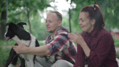 Gülümseyen bir adamın yaz parkında kadın ve köpeklerle oturup konuşması. Pozitif beyaz bir kocanın portresi. Eşi ve evcil hayvanlarıyla sabah keyfi yapıyor..