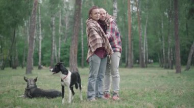 Parkta battaniyeyle örtülü duran ve konuşan beyaz bir çiftin geniş açılı görüntüsü. Sahiplerinin yanında oturan iki köpek. Mutlu erkek ve kadın dışarıda yaz sabahının tadını çıkarıyorlar..