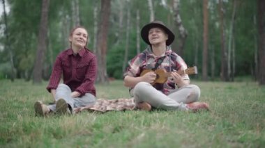 Şapkalı bir adam yaz parkında kız arkadaşı ya da karısı için ukulele çalarken önünde bulanık bir köpek vardı. Rahatlamış mutlu beyaz çift portresi. Evcil hayvanlarla battaniyede oturuyorlar..