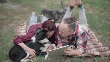 Rahatlamış çiftlerin ve evcil hayvanların tablet kullanarak yazlık çimlerde yatışının üst açısı. Beyaz bir kadın ve erkek köpeklerle dışarıda vakit geçirmenin keyfini çıkarıyorlar..