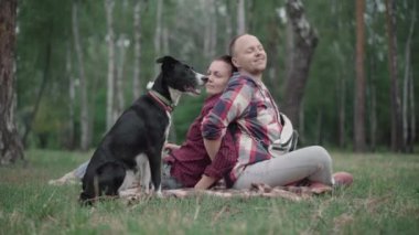 Ormandaki yaz çimlerinde sırt sırta oturan sevecen bir çiftle siyah beyaz bir köpek resmi. Olumlu gülümseyen beyaz erkek ve kadın evcil hayvanlarla parkta eğleniyorlar..