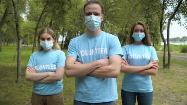 Vážní dobrovolníci v koronavirových obličejových maskách stáli se zkříženýma rukama a dívali se do kamery. Portrét skupiny mladých bělochů pózujících v letním parku. Covid-19 a ochrana životního prostředí — Stock video