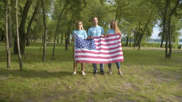 Grupp av leende volontärer poserar med amerikansk flagga på solig dag efter rengöring park. Porträtt av glad vit man och kvinnor står på tydlig äng och tittar på kameran. — Stockvideo