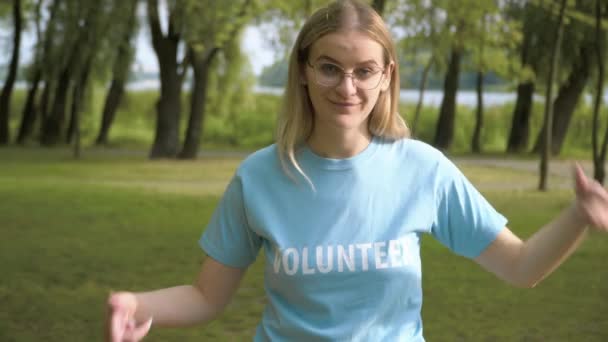 Retrato de uma jovem sorridente apontando para a palavra Voluntária na camiseta e olhando para a câmera. Senhora caucasiana confiante posando na luz solar depois de limpar o parque do lixo. Ecologia e ambiente. — Vídeo de Stock