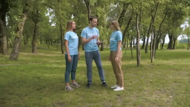 Amplio tiro de voluntarios jóvenes caucásicos deliberados discutiendo limpieza en el parque de verano. Tres hombres y mujeres seguros cuidando de la naturaleza. Ecología y protección medioambiental. — Vídeo de stock
