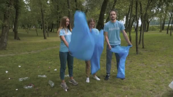 Voluntários confiantes abrem sacos de lixo e começam a limpar o parque no dia de verão. Retrato de tiro largo de homem e mulheres caucasianos sorridentes limpando prado no dia ensolarado. — Vídeo de Stock