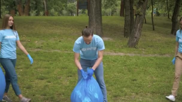Schlanke schöne kaukasische Frauen gehen zu einem Mann, der im Sommerpark riesige Müllsäcke verschließt. Porträt von drei positiv selbstbewussten kaukasischen Freiwilligen, die Wiese vom Müll säubern. — Stockvideo