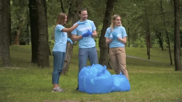 Широкий снимок положительных мужчин и женщин-добровольцев в защитных перчатках, стоящих с собранным мусором и разговаривающих. Портрет счастливой молодежи, очищающей летний парк от мусора. Экология и очистка — стоковое видео