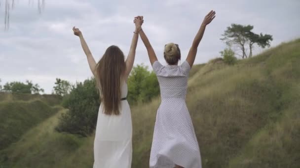 Вид сзади на двух стройных роскошных кавказских женщин, стоящих на улице в ветреный день в светло-белых платьях с поднятыми руками. Уверенная молодая мать и взрослая дочь наслаждаются отдыхом на природе. — стоковое видео