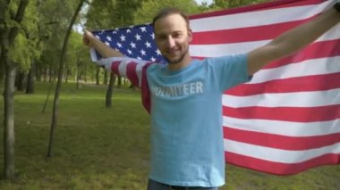 Güneşli yaz parkında Amerikan bayrağıyla poz veren pozitif erkek gönüllü. Beyaz tenli genç bir adamın portresi temizlikten sonra milli sembolle dönüyor ve hareket ediyor..