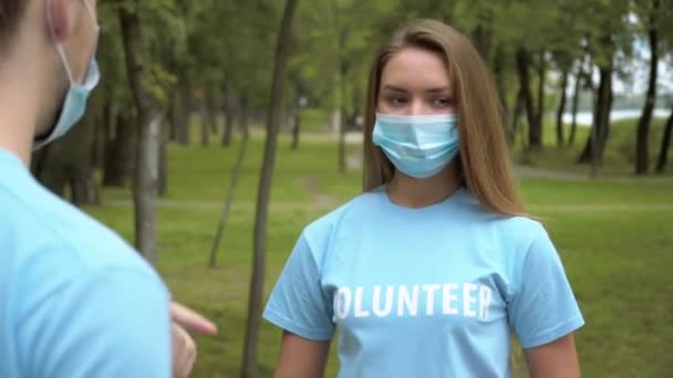 Une jeune femme positive portant un masque facial Covid et un T-shirt bénévole parlant avec un homme le jour de l'été à l'extérieur. Portrait de militants écologistes caucasiens confiants ayant un parc de nettoyage de rupture sur le verrouillage du coronavirus. — Video