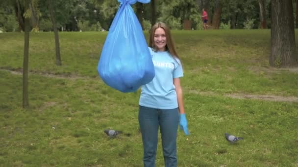 Šťastná dobrovolnice s obrovským pytlem na odpadky, usmívající se před kamerou a ukazující vítězné gesto. Portrét spokojené kavkazské mladé ženy pózující po úklidu parku venku. Ekologický koncept. — Stock video