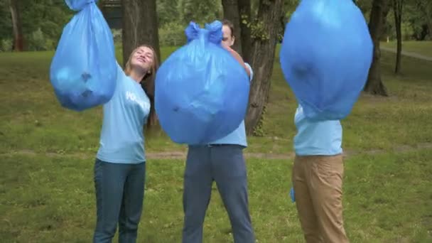 Tres voluntarios caucásicos levantando bolsas de basura y sonriendo. Retrato de felices eco-activistas orgullosos posando con basura recogida en el parque de verano. Contaminación ambiental y limpieza. — Vídeos de Stock