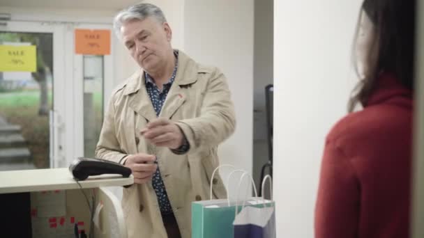 Hombre mayor de confianza que paga por la compra con tarjeta de crédito, toma bolsas de la compra y se va. Retrato de elegante comprador masculino caucásico utilizando el pago inalámbrico en la tienda. Comercio moderno. — Vídeo de stock