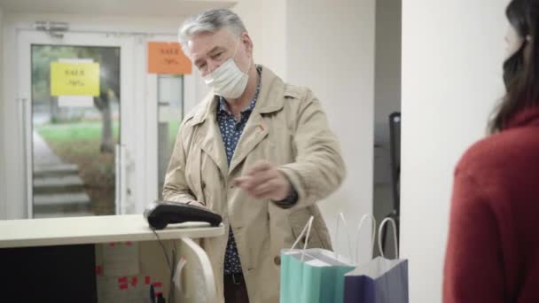 Hombre mayor en máscara facial Covid utilizando el pago sin contacto en la tienda. Retrato de cliente varón seguro pagando en línea con tarjeta de crédito y saliendo con bolsas de la compra. Concepto de pago inalámbrico. — Vídeos de Stock