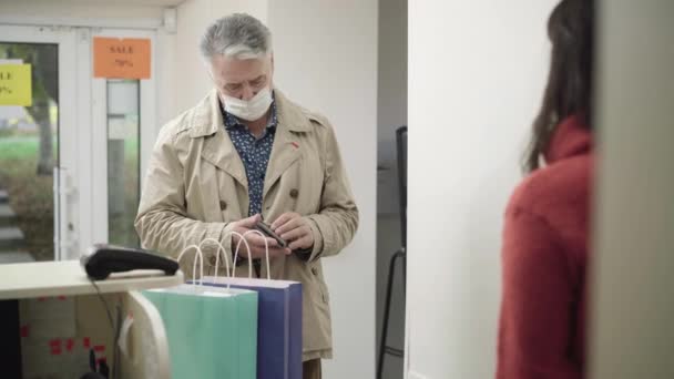 Portrait of senior Caucasian man in Covid-19 face mask looking in empty wallet in shop and leaving. Embarrassed male buyer walking away without purchase. Poverty during coronavirus pandemic. — Stock Video