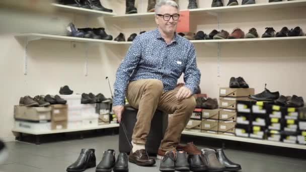 Stubborn senior man trying on too small boot in shoe store. Wide shot of elegant Caucasian male shopper in eyeglasses sighing and looking at footwear standing in front. Shopping and consumerism. — Stock Video