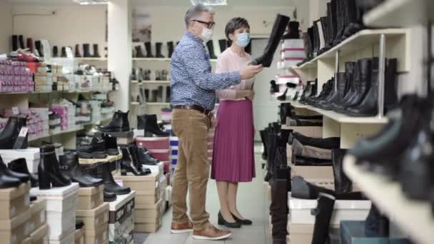 Covid-19のシニア白人男性と女性の幅広いショットは、コロナウイルスパンデミックの間に靴店で履物を選択する顔マスク。自信を持ってエレガントなカップル選択ブーツで店. — ストック動画
