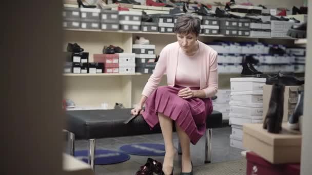 Breed schot van serieuze blanke senior vrouw zittend in de schoenenwinkel en hoge hakken aan het passen. Portret van een elegante vrouwelijke klant die schoeisel kiest in de winkel tijdens de verkoop op vrijdag. Winkelconcept. — Stockvideo