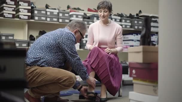 Flot senior kaukasisk mand hjælper elegant smuk kvinde til at sætte på høje hæle i skobutikken. Portræt af lykkelige par pensionister vælger fodtøj i butikken. Familiekøb koncept. – Stock-video