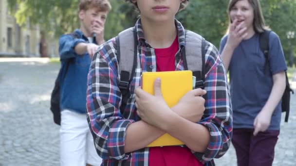 本を背景から笑っている同級生として屋外に立っている認識できないオタク白人の少年。いじめられた女子高生が校庭でいじめを受ける. — ストック動画