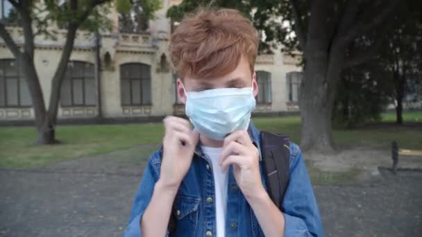 红头发的小男孩戴上Covid-19型面罩，看着摄像机。考拉韦病毒大流行期间，白人学童在户外摆姿势的画像。Covid生活方式概念. — 图库视频影像