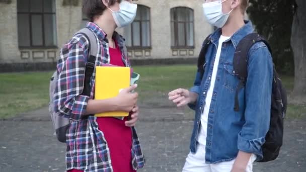 Due ragazzi irriconoscibili in maschere facciali Covid-19 che parlano durante una pausa nel cortile della scuola. Studenti caucasici che chiacchierano durante l'epidemia di pandemia di coronavirus dopo aver studiato. Assistenza sanitaria e istruzione. — Video Stock