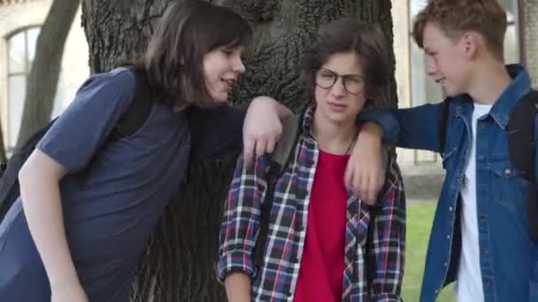Compañeros bromeando nerd chico caucásico en el patio de la escuela. Retrato de morena asustada colegial en gafas cerrando orejas. Concepto de violencia e intimidación. — Vídeos de Stock
