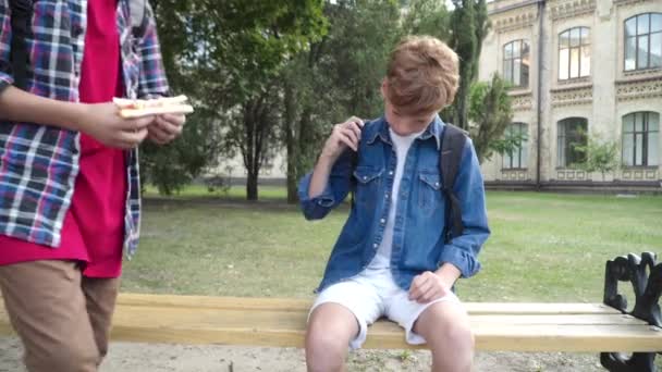 Bruneta Kavkazský chlapec sdílení sendvič s zrzavý přítel sedí na lavičce na školním dvoře. Portrét malých kamarádů, kteří mají přestávku po studiu. Koncept přátelství a štědrosti. — Stock video