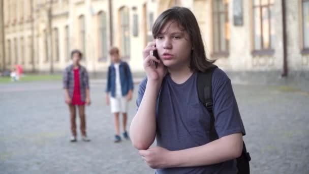 Nadváha Kavkazský školák mluví na smartphonu a utíká od agresivních spolužáků kráčejících v pozadí. Portrét vyděšeného šikanovaného chlapce odcházejícího. Koncept šikany a strachu. — Stock video
