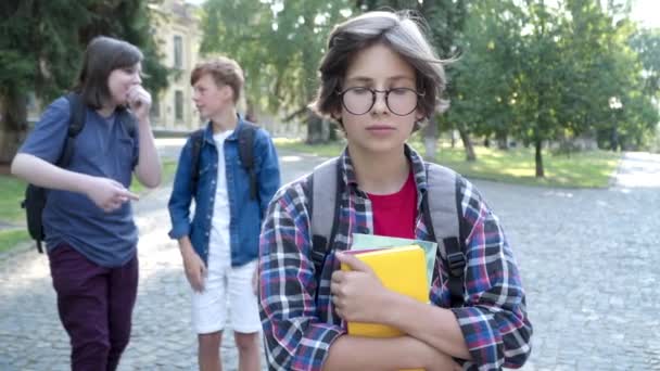 Geniale schooljongen in brillen die op het schoolplein staan als wazige klasgenoten die lachen en naar hem wijzen vanaf de achtergrond. Portret van gepest intelligente blanke jongen buiten. — Stockvideo